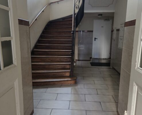 Treppenhaus mit Holztreppe, schmiedeeisernem Geländer und frisch gewischtem Fliesenboden