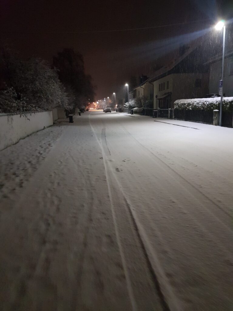 Winterdienst: Straße mit frischem Neuschnee bei Nacht, Straßenlaternen und verschneite Hecken